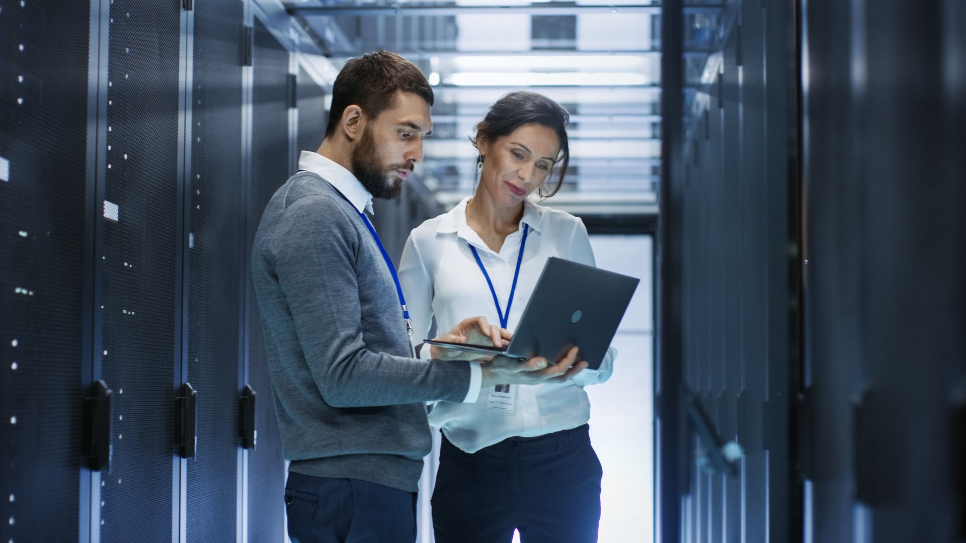 Male IT Specialist Holds Laptop and Discusses Work with Female Server Technician. They're Standing in Data Center, Rack Server Cabinet is Open.